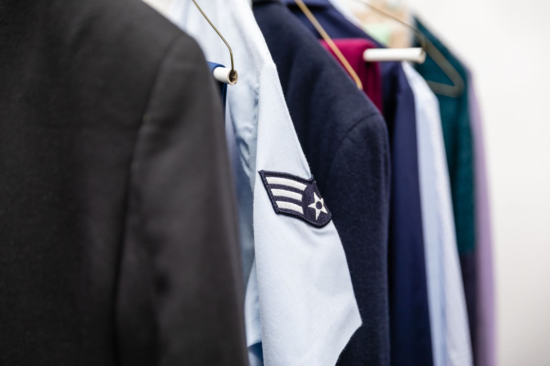 A row of clothing on hangers, including a black blazer and a light blue shirt with a military-style patch.