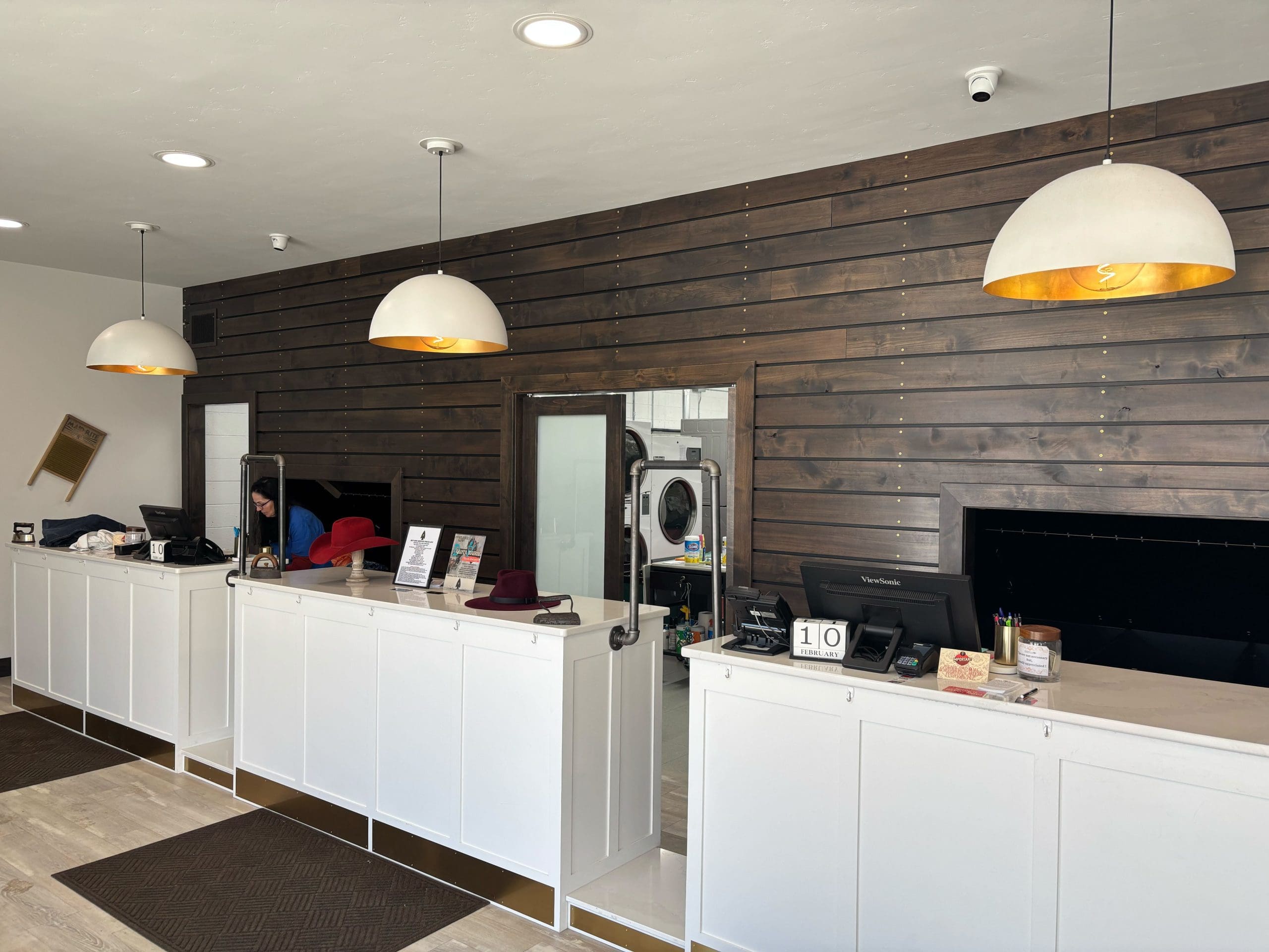 A modern dry cleaning counter with white cabinets and a wooden wall. Clothing items are visible behind the counter. Two pendant lights hang above.