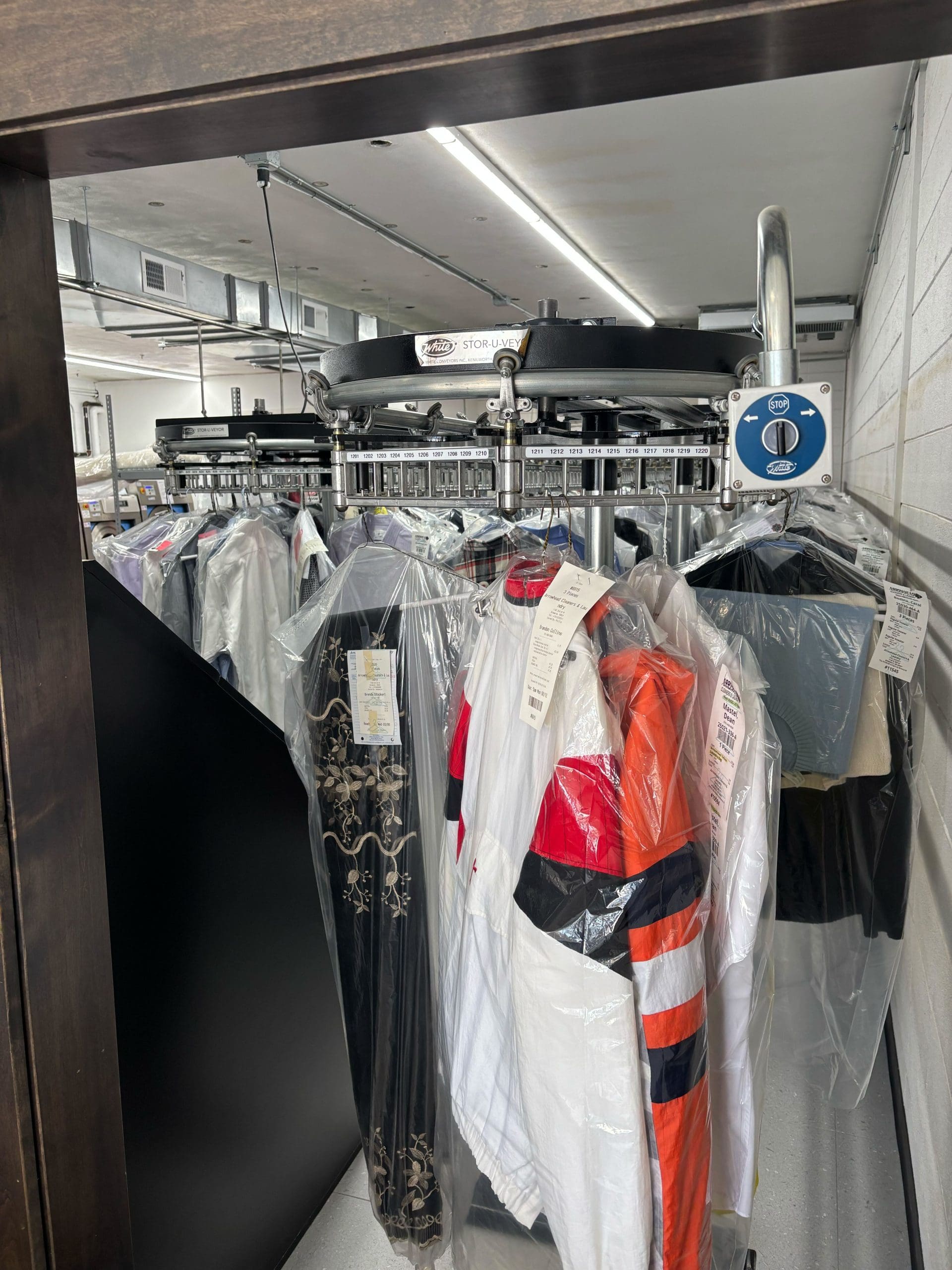 Clothes on hangers are covered in plastic, hanging from a conveyor system in a dry cleaning facility.