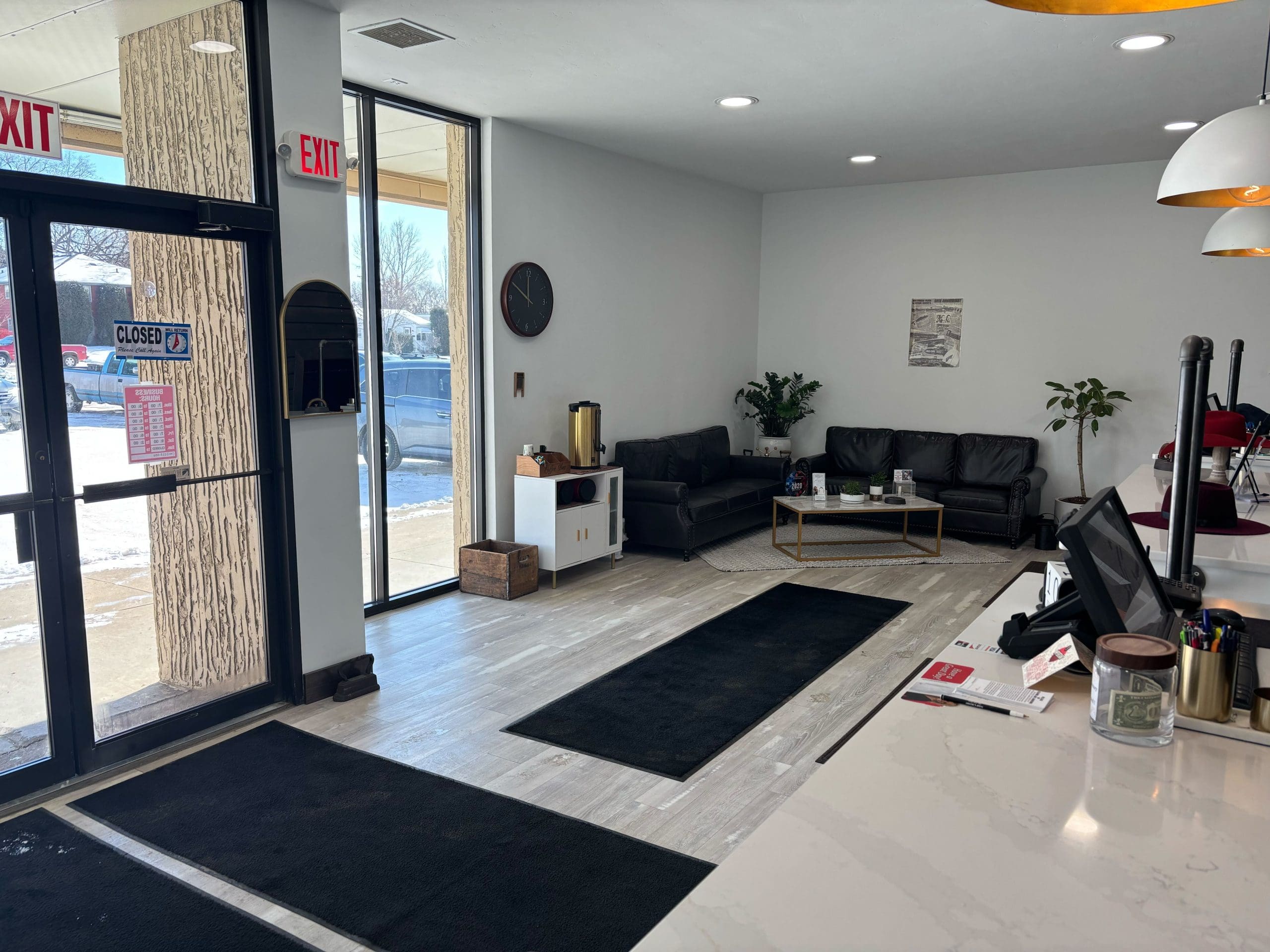 Office lobby with a reception desk, black sofas, a wall clock, potted plants, and large windows. A 