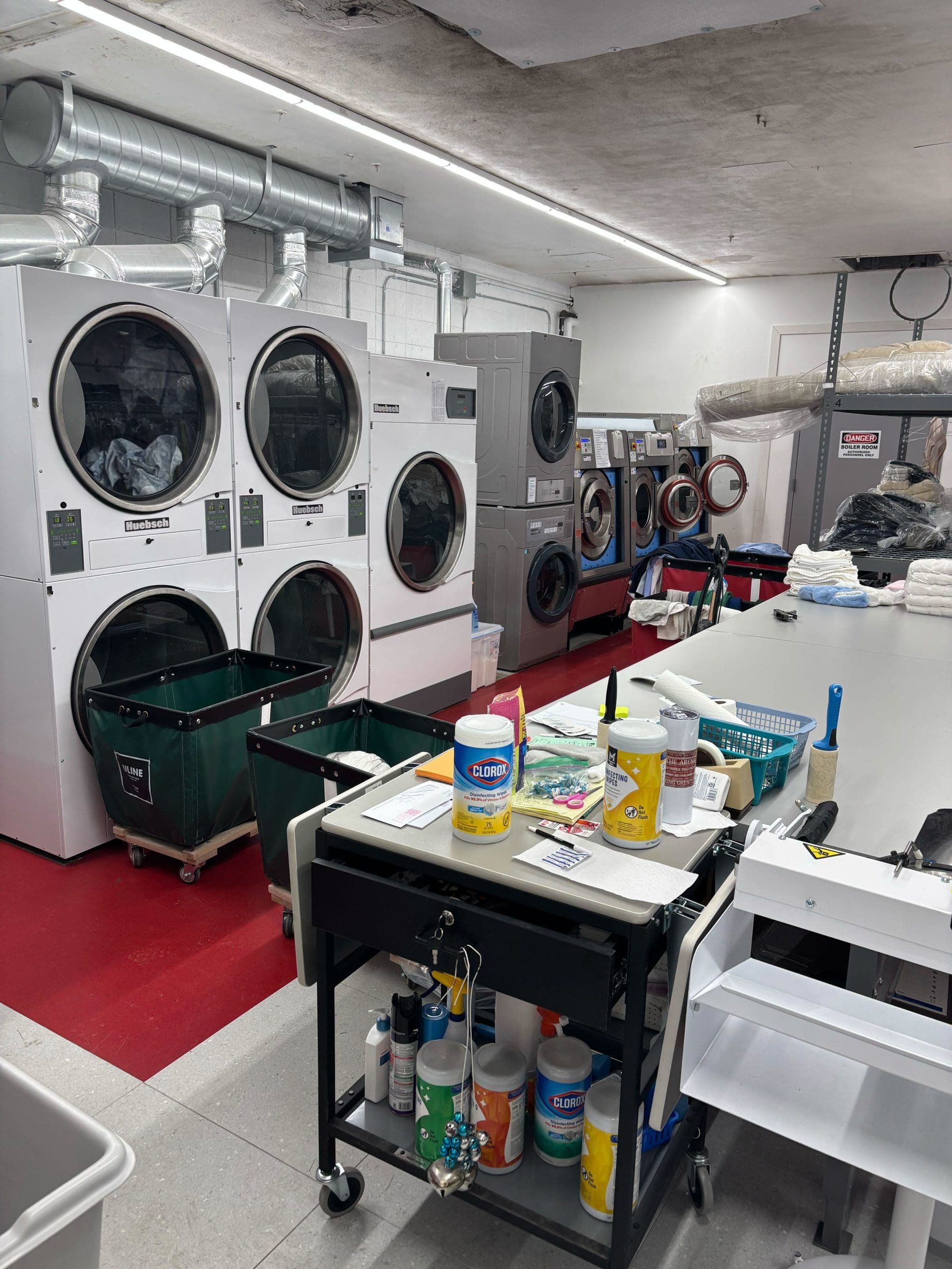 Commercial laundry room with washing machines, dryers, cleaning supplies on a cart, and folding tables. The floor is red and white, and ductwork is visible on the ceiling.