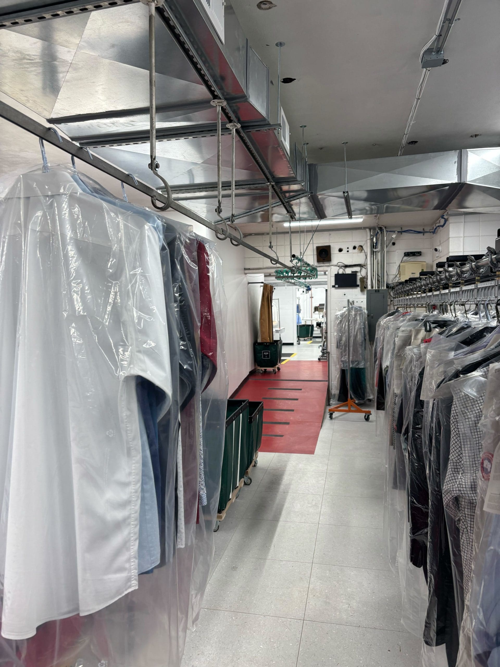 A dry cleaning facility with shirts and garments wrapped in plastic hanging on a conveyor system. Rows of clothing are visible, with an industrial setting in the background.