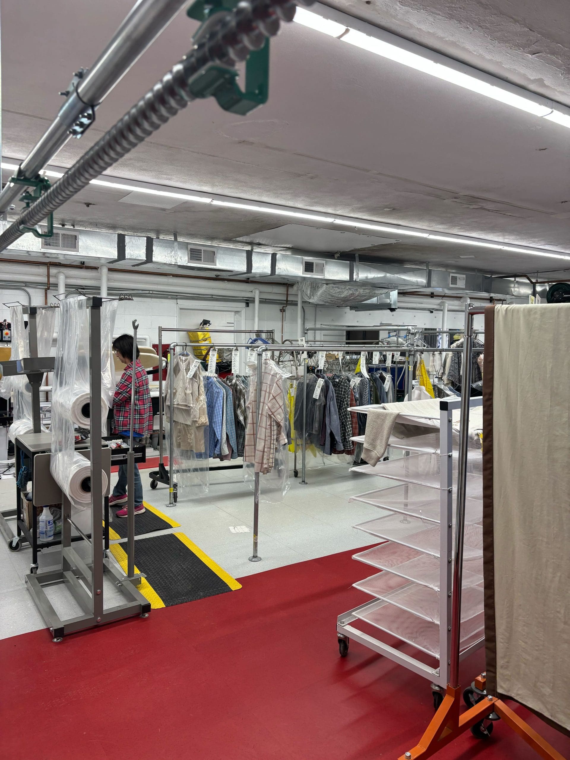 Industrial laundry room with clothing racks, garment bags, and a worker using equipment. Red and white flooring, metal pipes overhead, and clothes hanging organized on racks.