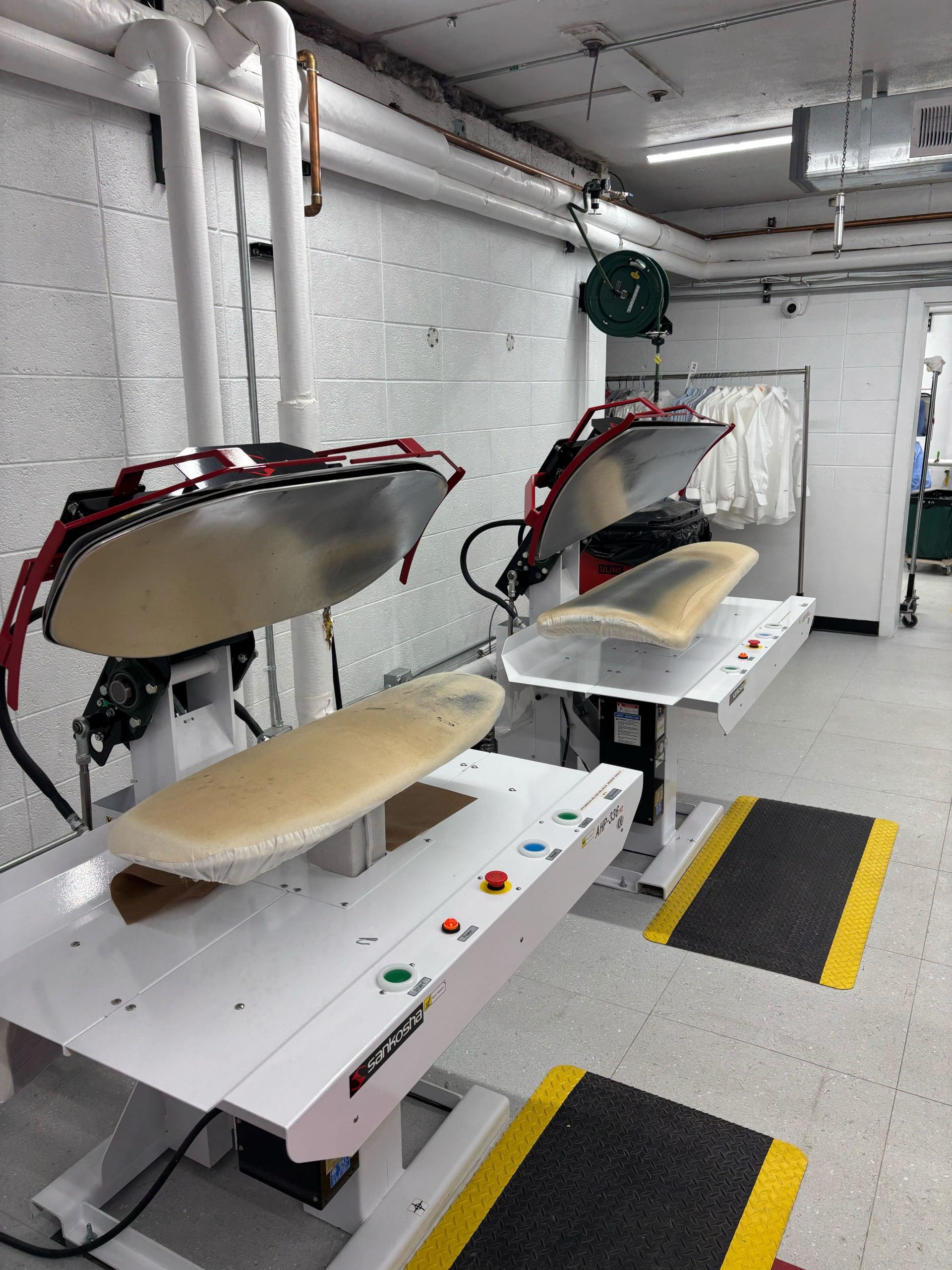 Two industrial ironing machines in a laundry facility, with white lab coats hanging in the background.