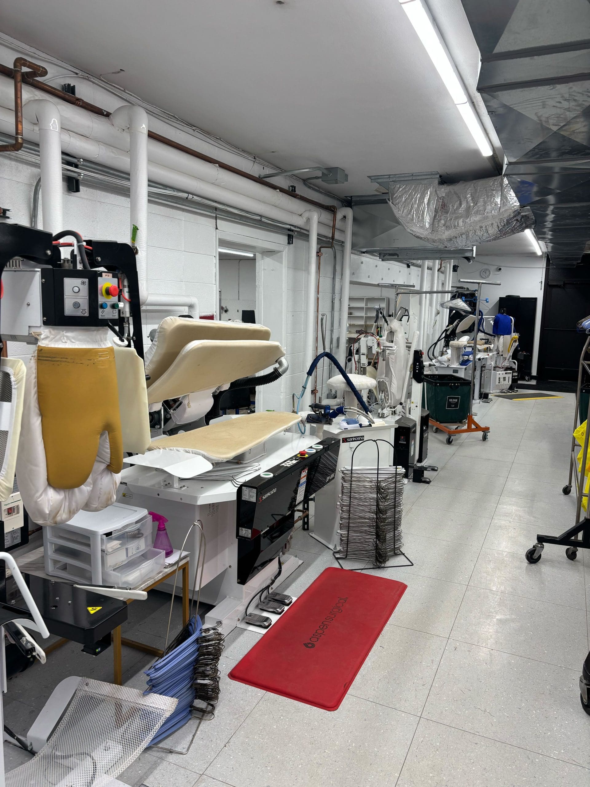 A laboratory-like room features multiple workstations with dental or medical equipment, including chairs, hoses, and various tools. The space is organized and has overhead lighting.