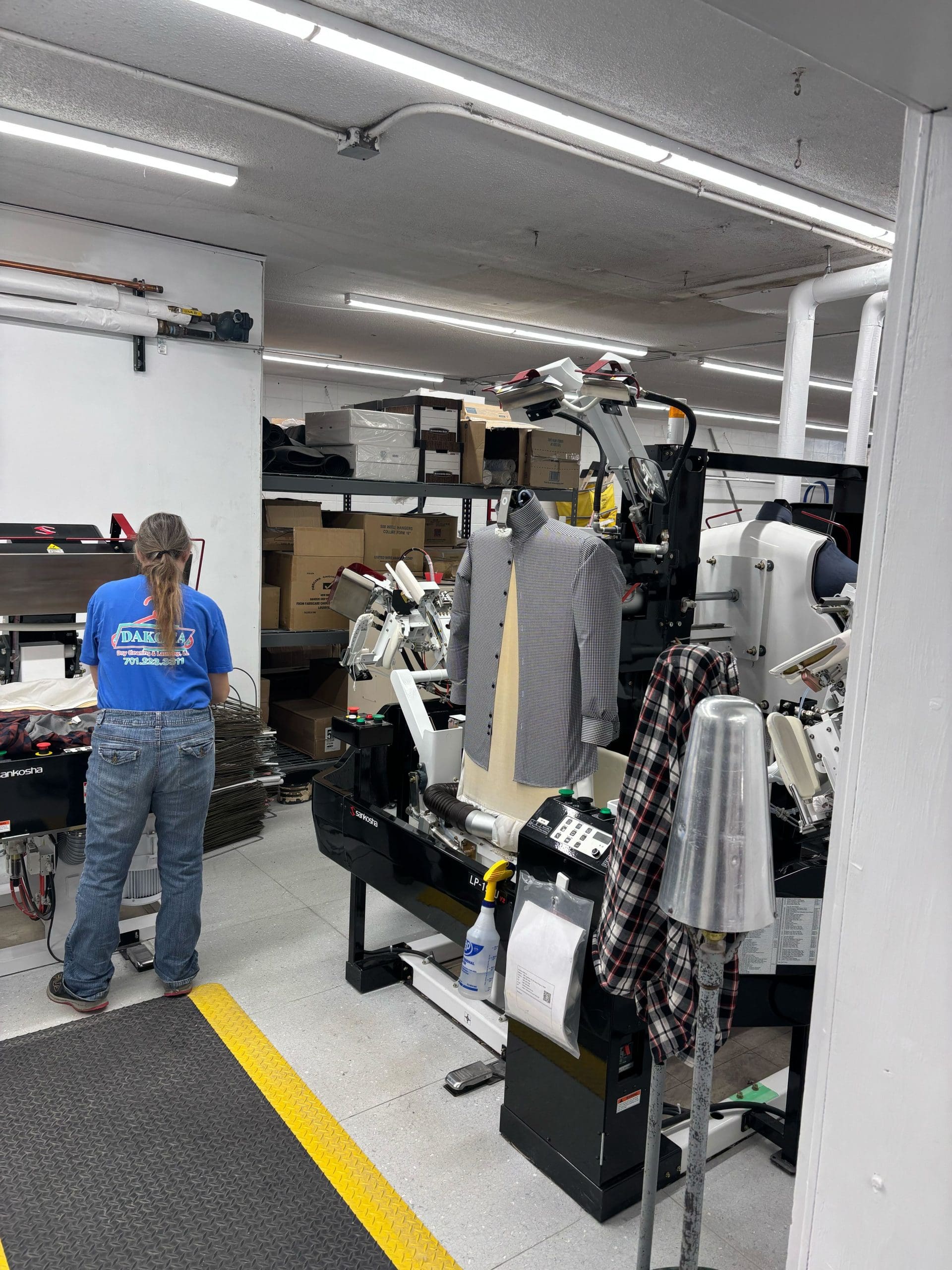 Person operating a garment pressing machine in a workshop. A suit jacket is placed on the machine, and boxes are stacked in the background.
