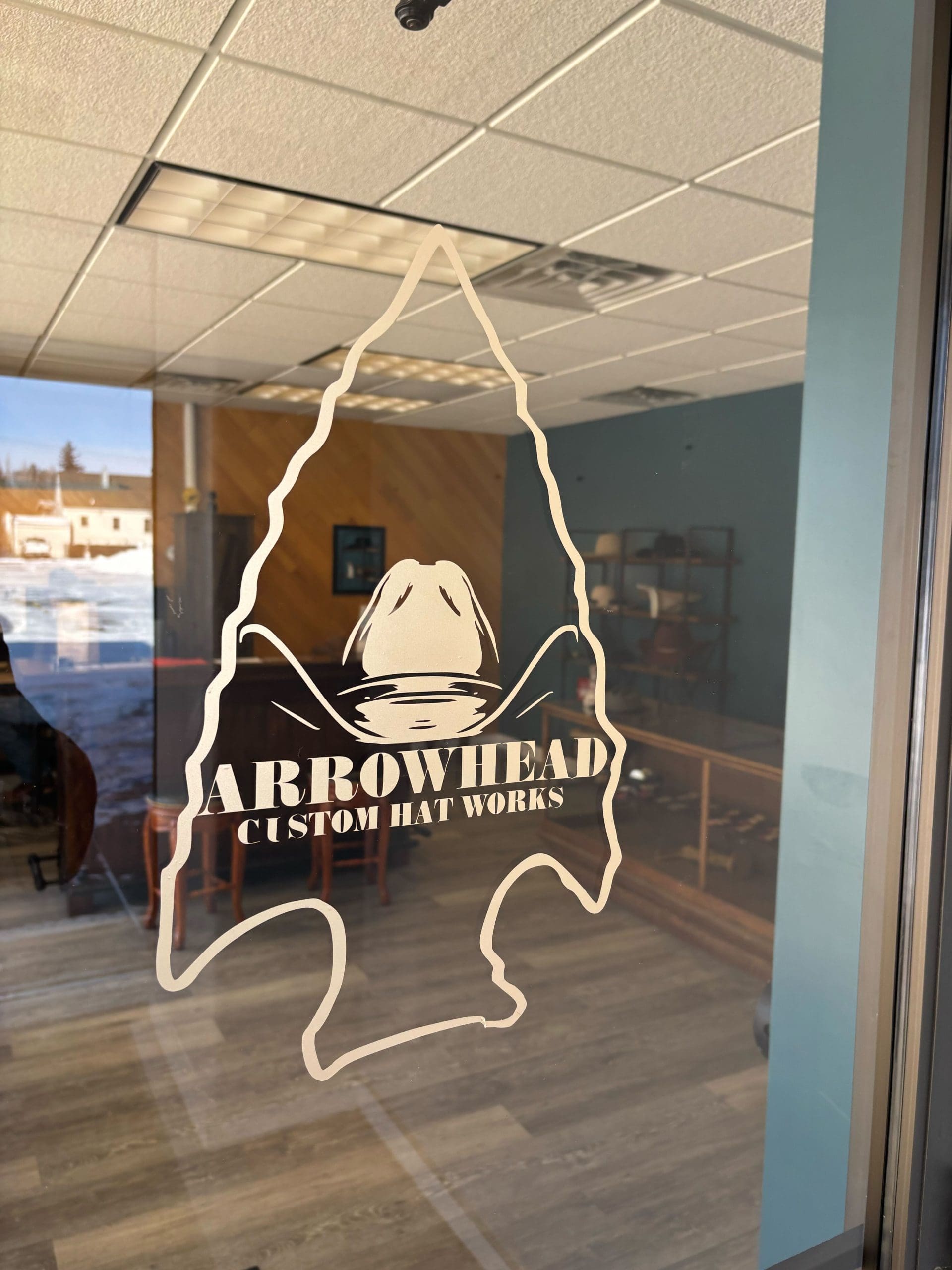 Arrowhead Custom Hat Works logo on glass door, featuring an arrowhead outline with a hat inside. Interior space visible through glass.