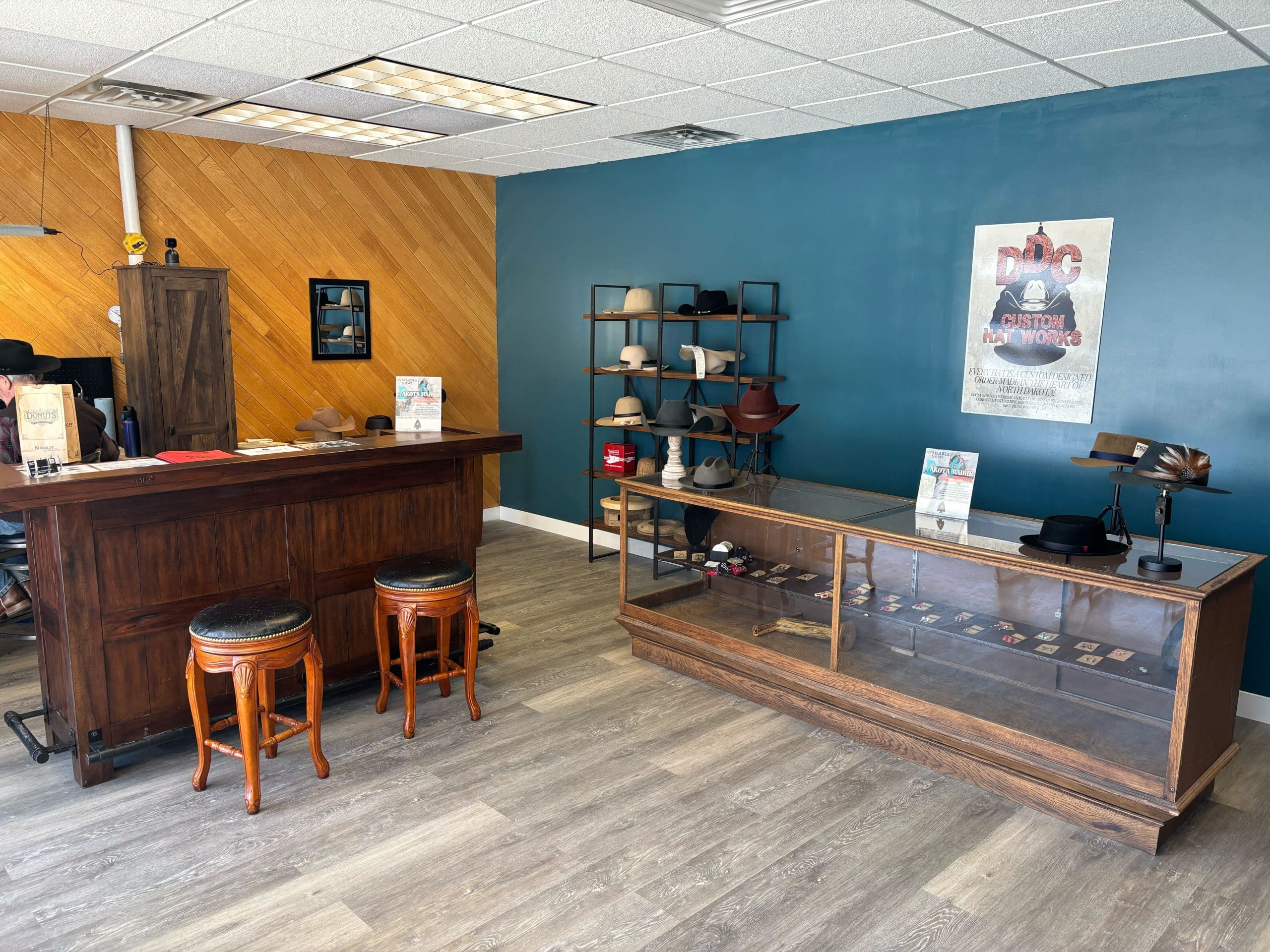 A hat shop interior with two stools at a wooden counter, a glass display case with hats, and a wall shelf holding various hats. Wood and blue walls create a stylish setting.