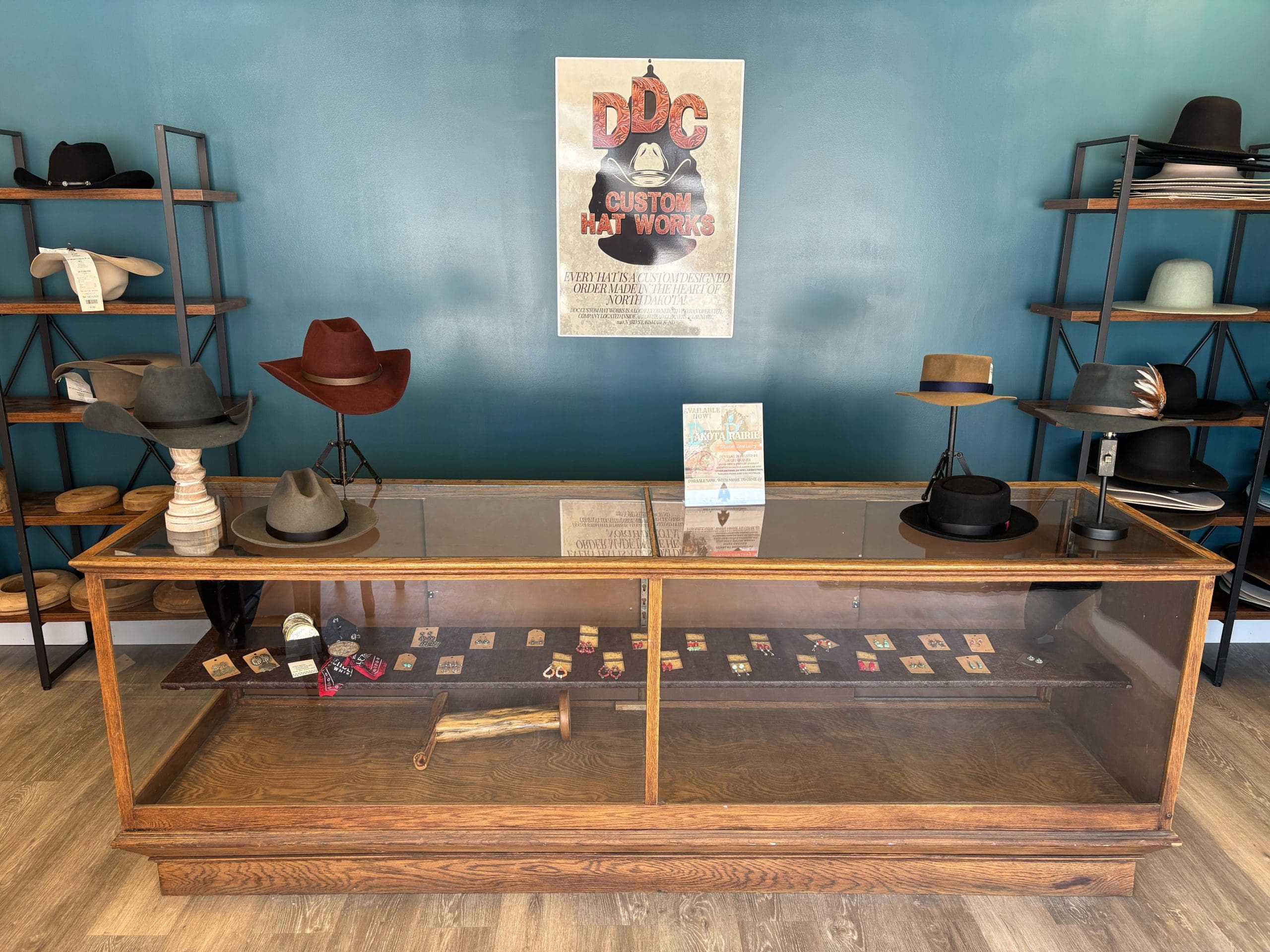 A hat shop display featuring various hats on and around a vintage glass counter. The backdrop is a teal wall with a poster and shelves holding hats and related items.