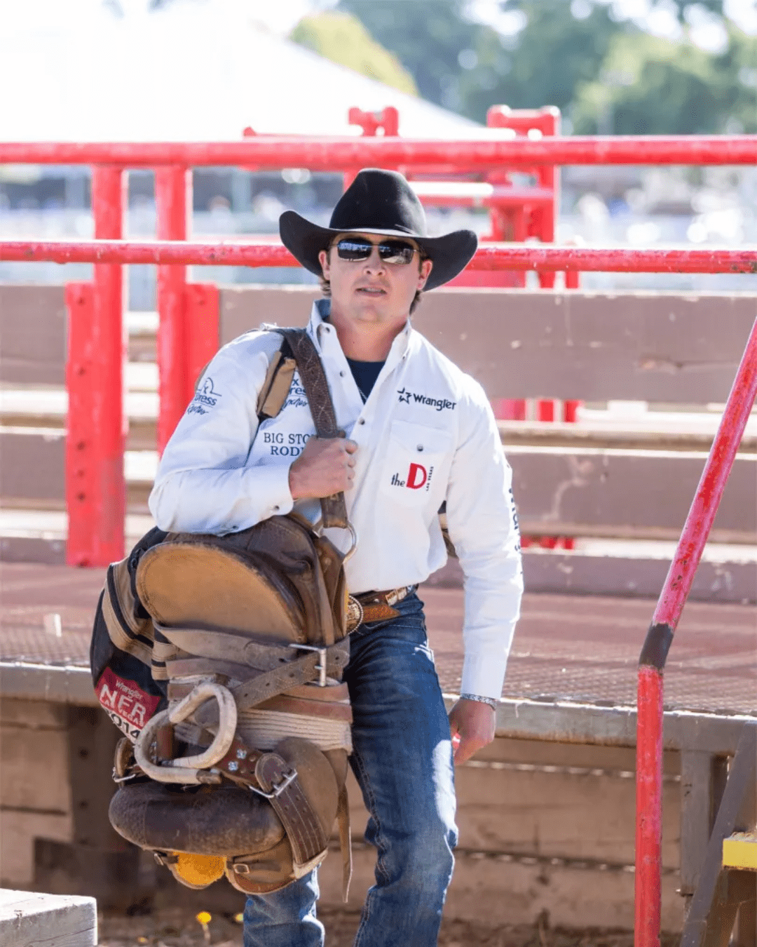 Cowboy in a black hat and sunglasses carries a saddle, wearing a white shirt with logos, blue jeans, and a brown belt. Red and brown railing in the background.