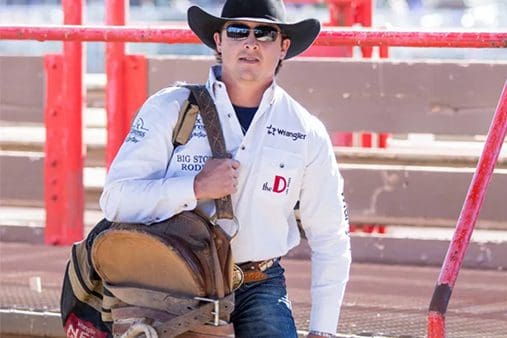 A person in a cowboy hat and sunglasses stands near a rodeo arena, holding a saddle, wearing a white shirt with logos.