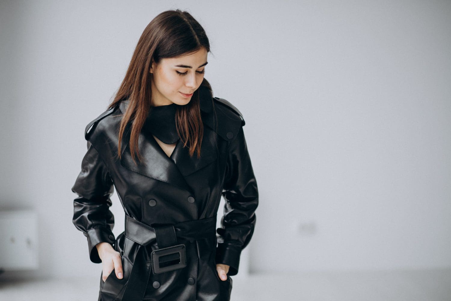 Woman in a black leather trench coat stands with hands in pockets, looking down, against a plain background.