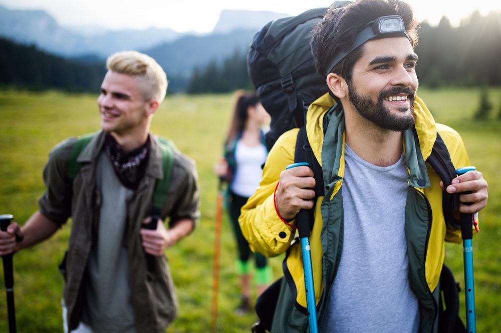 Two men with backpacks and hiking poles smile while walking in a grassy outdoor setting, with mountains in the background.