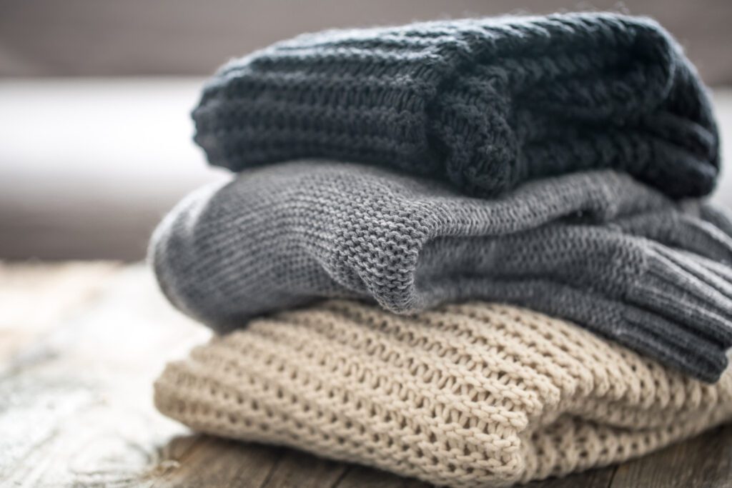 Three folded knitted sweaters in dark gray, light gray, and beige colors stacked on a wooden surface.