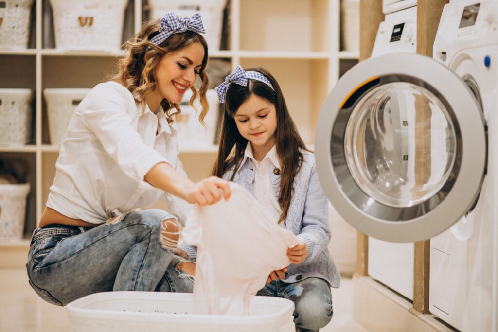 An adult and a child load laundry into a washing machine together in a laundry room, both wearing casual clothes and headbands.