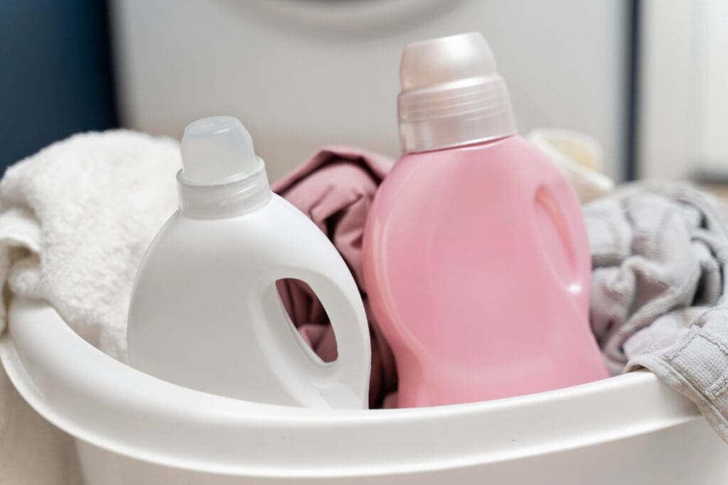 A white plastic basket contains two bottles of laundry detergent, one white and one pink, along with assorted laundry items.