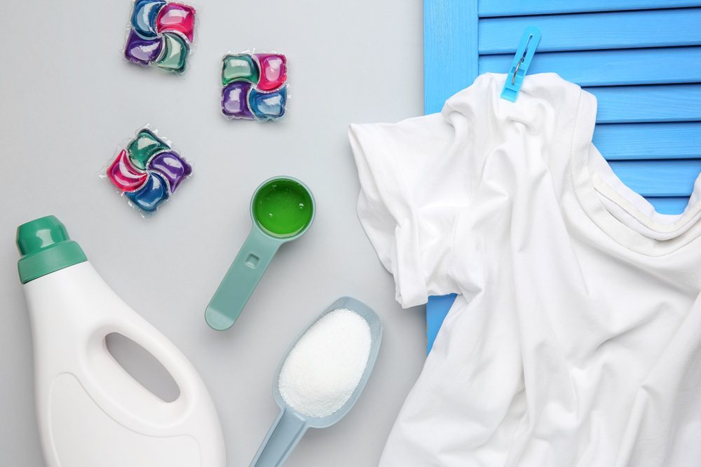 Laundry detergent bottle, a scoop of powder, a measuring cup with liquid, laundry pods, and a white t-shirt on a blue background.