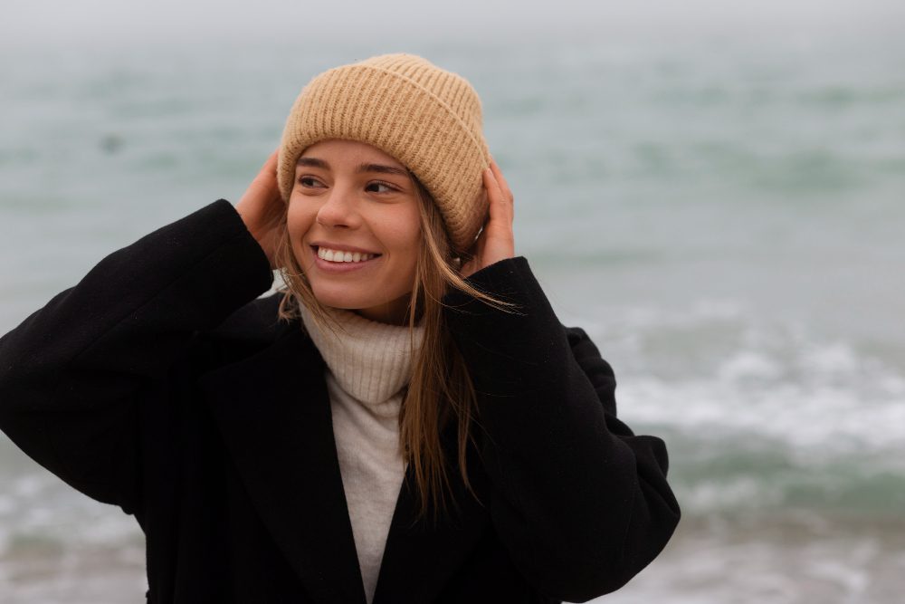 Woman wearing a tan knit beanie, black coat, and light turtleneck stands near the ocean, smiling and holding her hat with both hands. The background is slightly foggy.