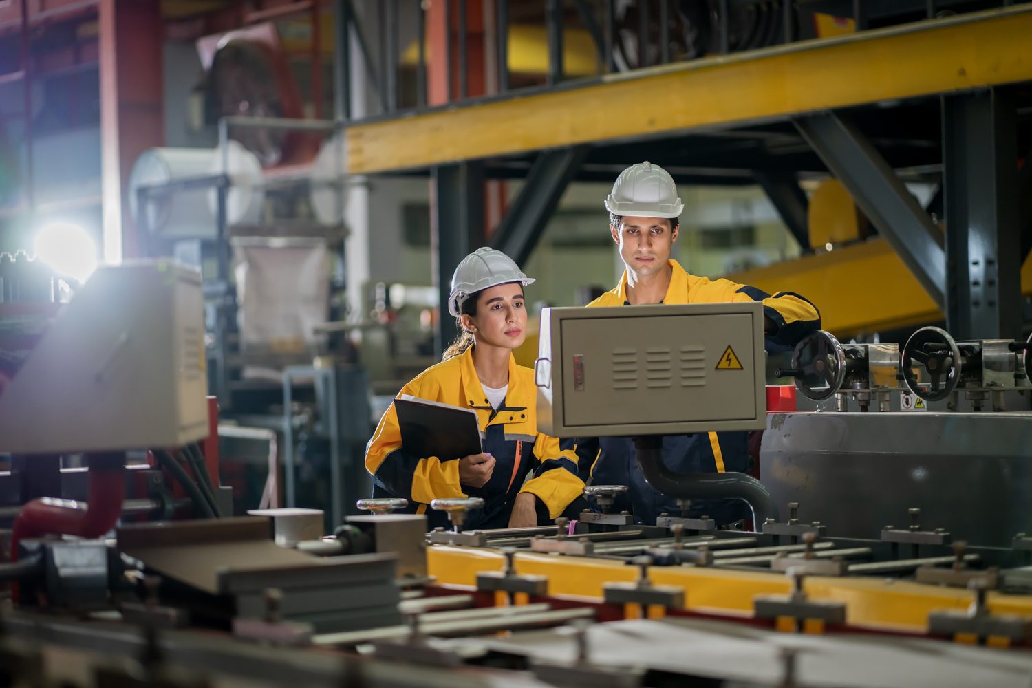 Two workers wearing hard hats and yellow safety jackets stand in a factory, inspecting machinery and holding a clipboard. Industrial equipment and shelves are visible in the background.