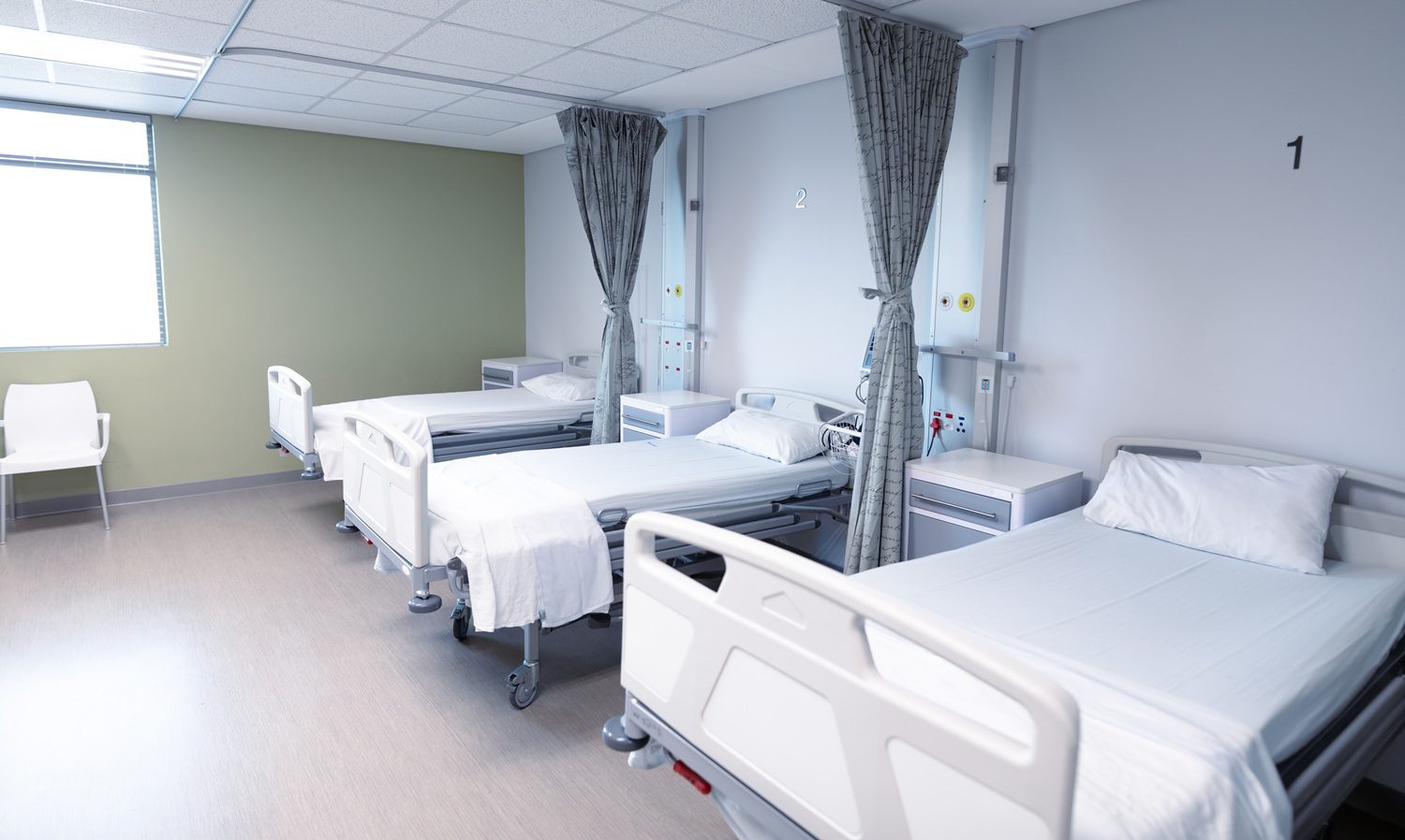 Three empty hospital beds with white linens are arranged in a row in a clean, modern hospital room, separated by privacy curtains.