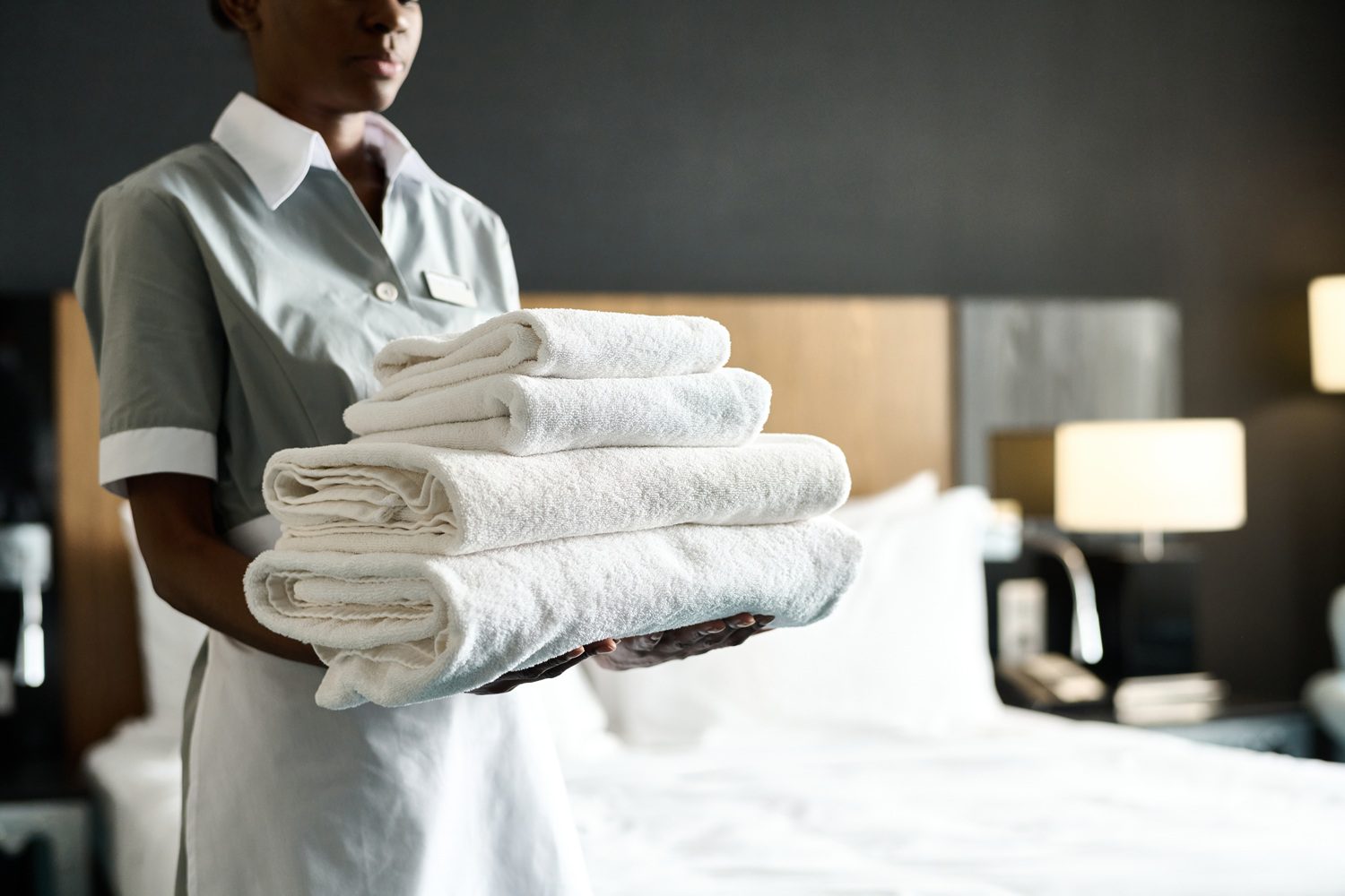 A hotel staff member in uniform holds a stack of clean white towels in a hotel room with a made bed and bedside lamps.