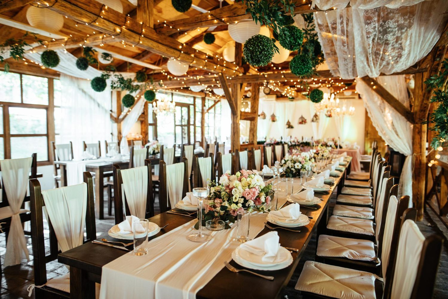 Long banquet tables are set for a formal event in a rustic, wooden venue decorated with string lights, white drapes, greenery, and floral centerpieces.