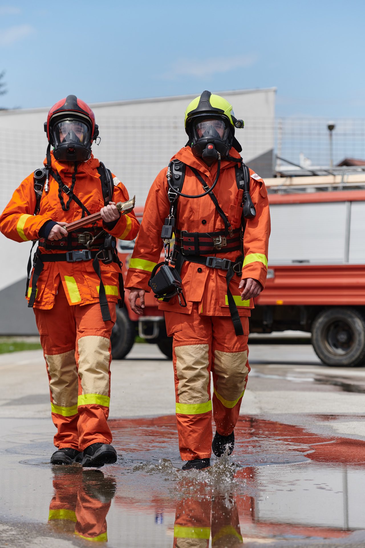 Two firefighters in full protective gear and masks walk on wet pavement near a fire truck.