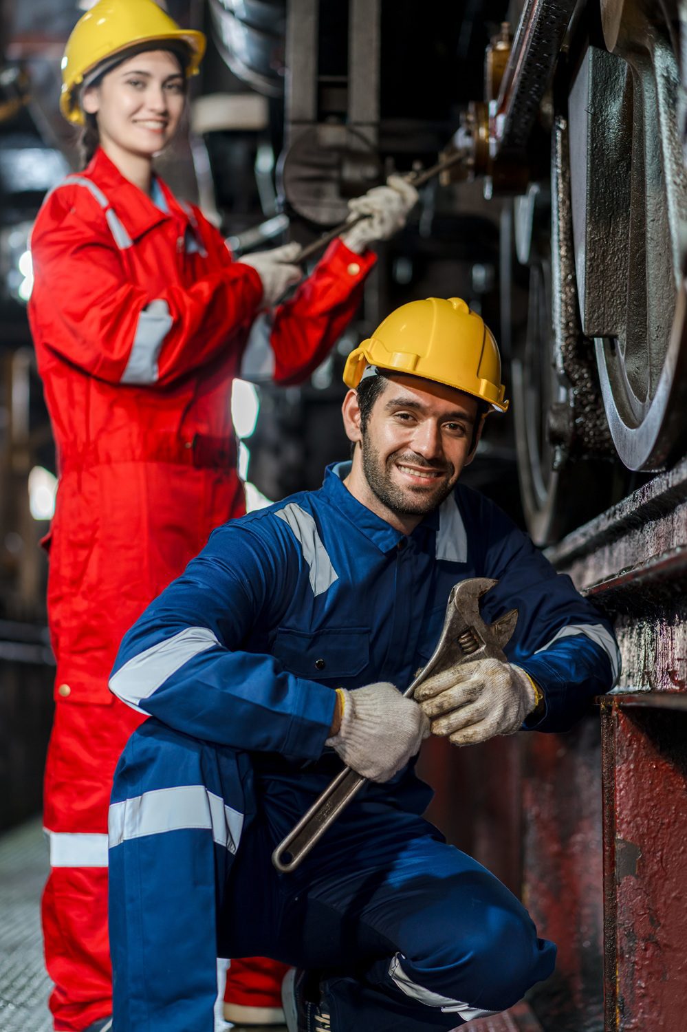 Two industrial workers wearing safety helmets and coveralls work on machinery; one kneels holding a wrench, while the other stands in the background adjusting equipment.