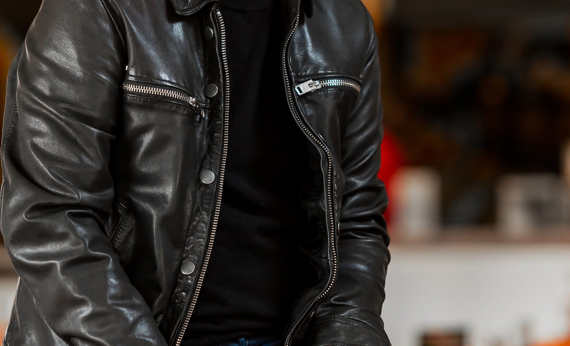 Person wearing a black leather jacket and black shirt, seated indoors with a blurred background.