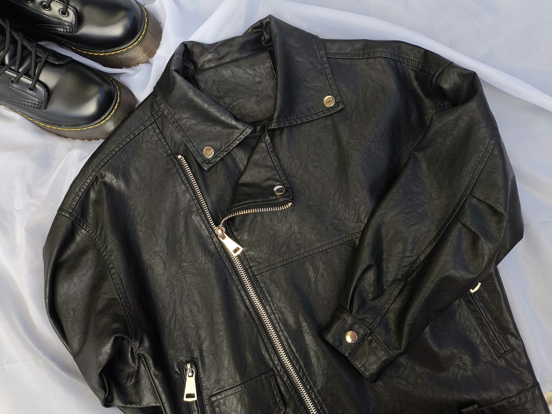 A black faux leather jacket with silver zippers and snap buttons, laid flat next to a pair of black lace-up boots on a light fabric background.