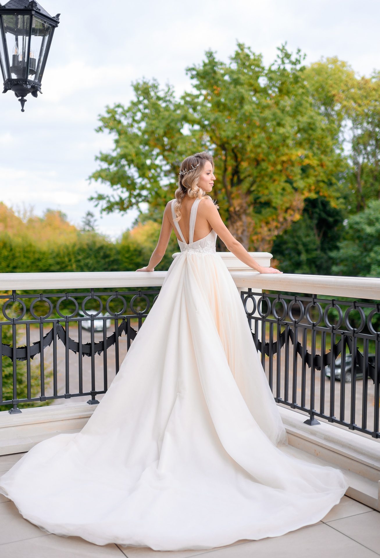 A woman in a white, floor-length wedding dress stands on a balcony, facing away, with trees and a lamp post visible in the background.