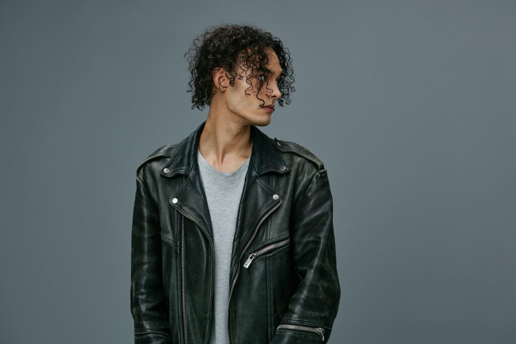 A person with curly hair wearing a black leather jacket and gray shirt stands against a plain gray background, looking to the side.