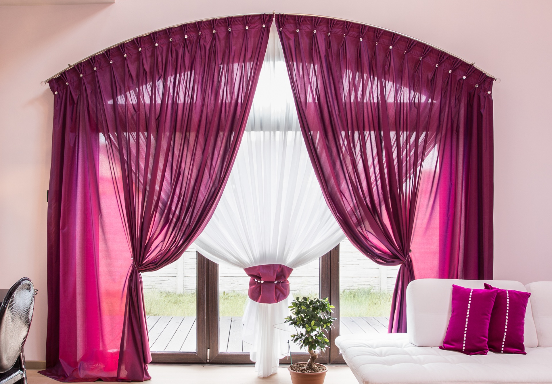 Purple sheer curtains are draped and tied back over white curtains on a large arched window; a potted plant and white sofa with purple pillows are in the foreground.