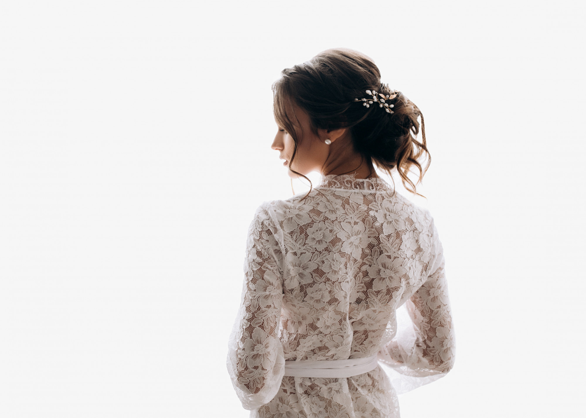 Woman in a white lace dress with an updo hairstyle, decorated with pearl hairpins, standing against a white background, viewed from behind and slightly to the side.