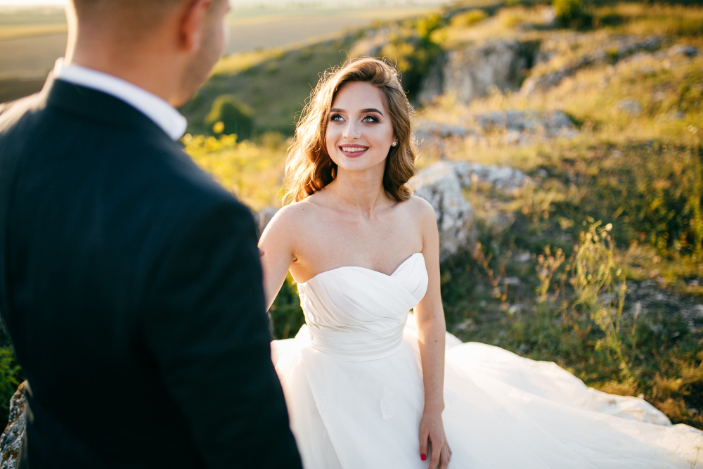 A bride in a strapless white gown stands outdoors on rocky terrain, smiling at a man in a dark suit facing her.
