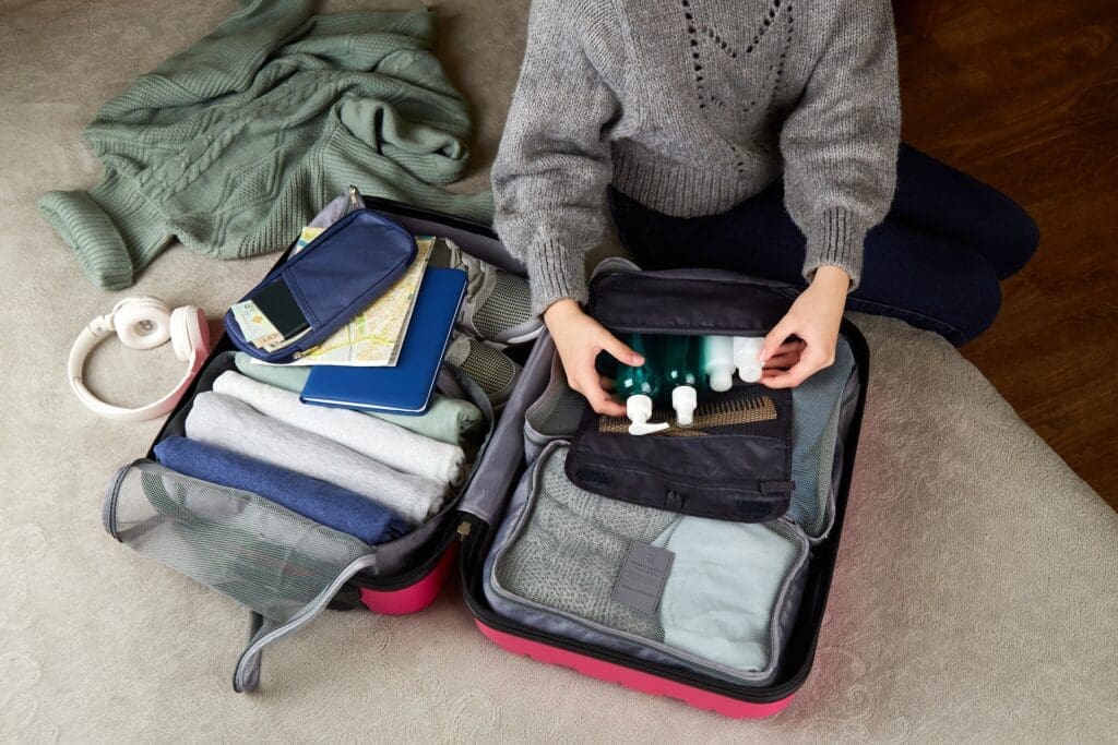 Person sitting on a bed packs a suitcase with neatly folded clothes, toiletries, travel documents, and a map. A sweater and headphones are placed nearby.