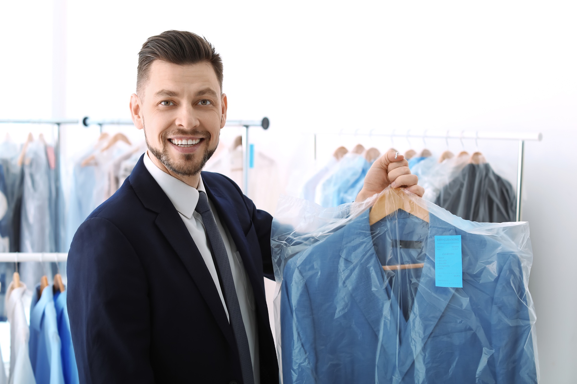 A man in a suit smiles while holding a freshly dry-cleaned jacket covered in plastic at a dry cleaning shop, with other clothes on hangers in the background.