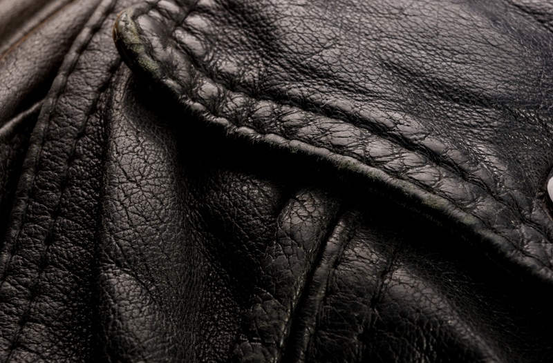 Close-up of a black leather jacket showing textured, wrinkled material, visible stitching, and part of a pocket or flap.