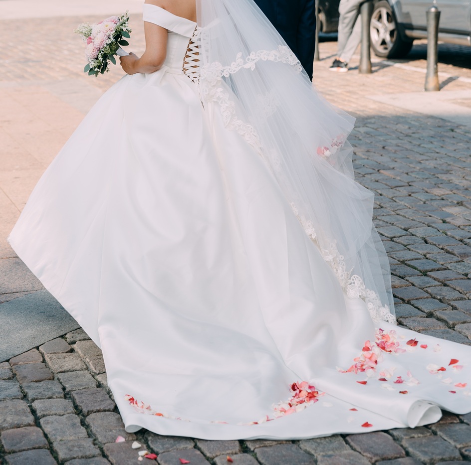 A bride in a white dress holds a bouquet, with pink flower petals scattered on her train, standing on a cobblestone street.