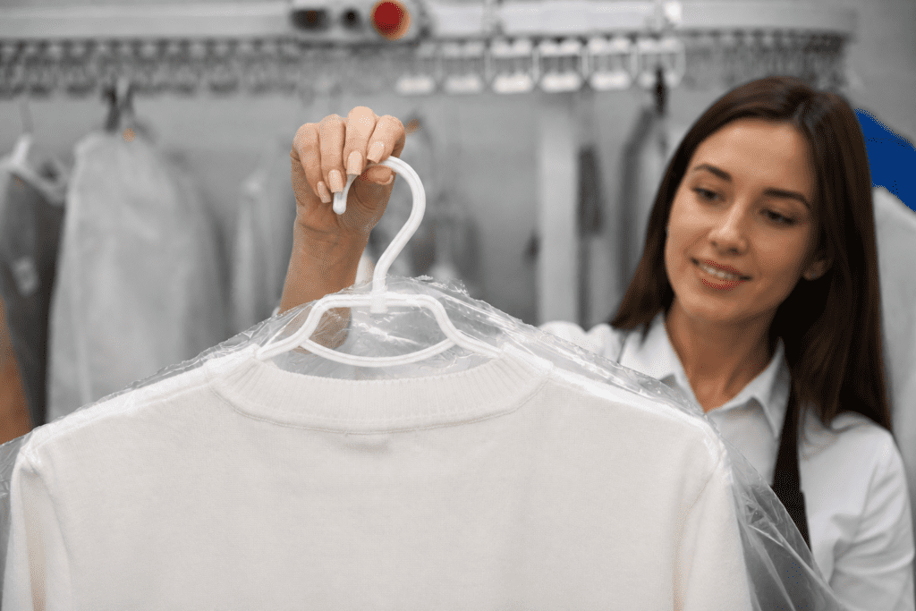 A woman holds a hanger with a white sweater covered in plastic at a dry cleaning facility, with clothes hanging in the background.